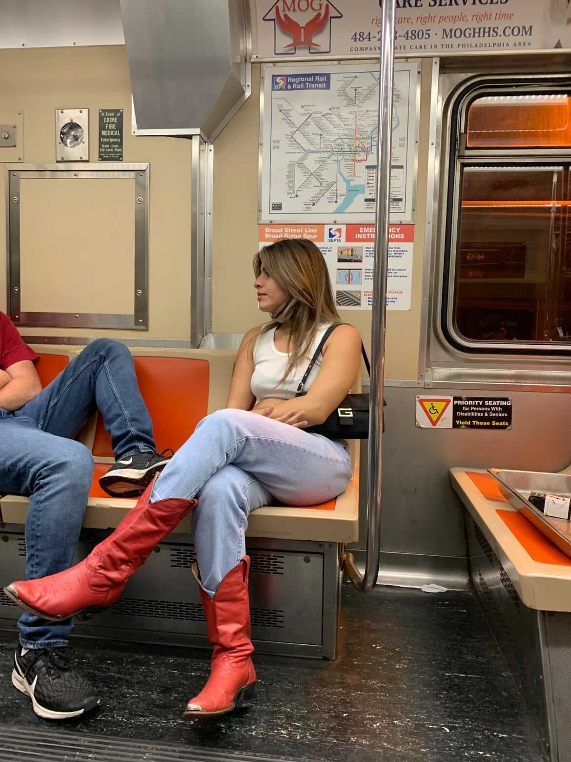 A girl sitting on the NYC subway she is wearing a white tank top, blue jeans, red cowboy boots, and a vintage guess handbag.