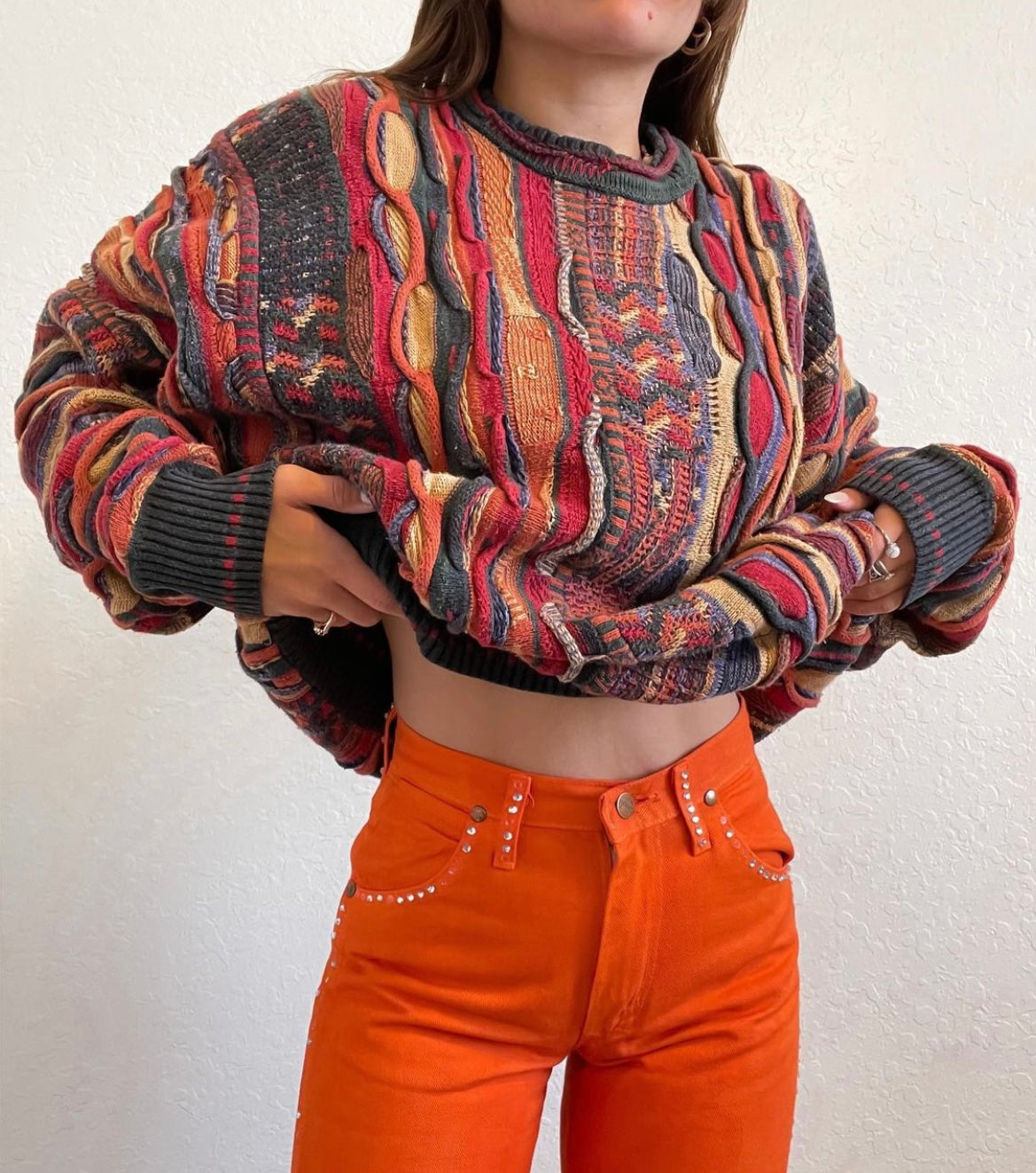 A model wearing a vintage coogi sweater with orange, red, yellow, and gray detailing. The model is also wearing vintage orange wranglers pants.